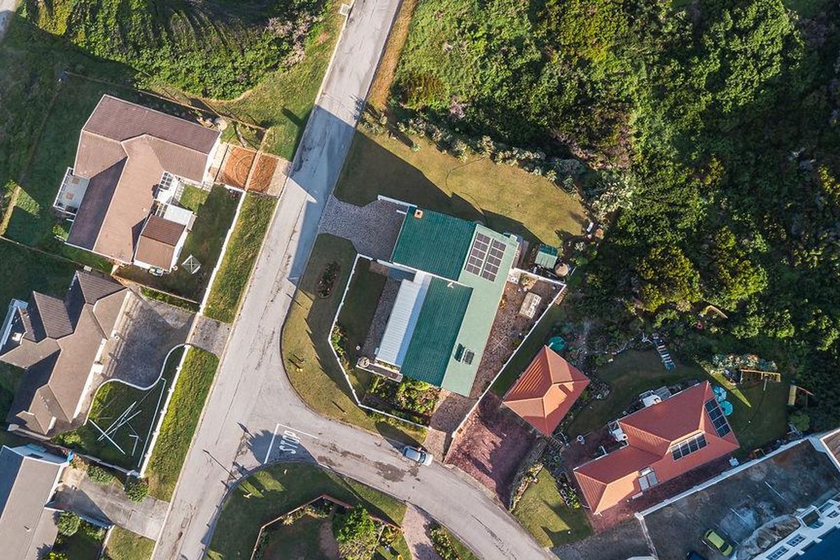 Aerial view of a residential property featuring a green pitched roof with solar panels, a paved driveway, and surrounding landscaped garden areas.