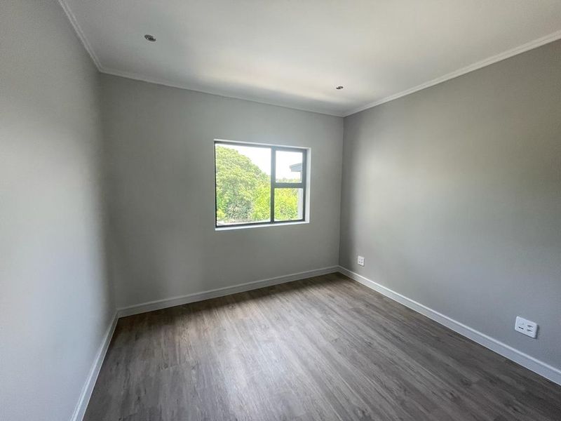 A bright, empty bedroom featuring modern grey laminate flooring, neutral walls, and an aluminium-framed window with a pleasant garden view.
