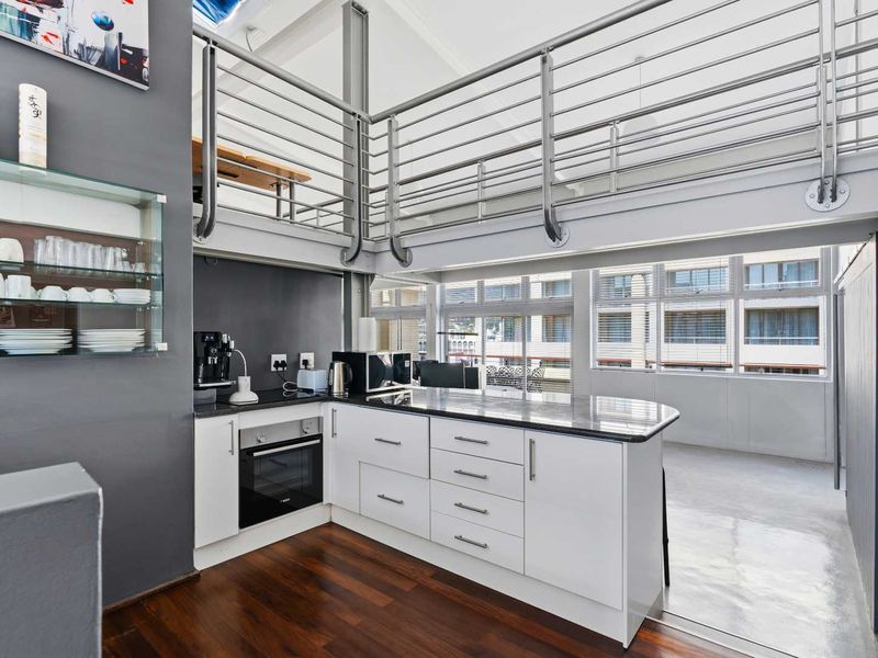 Modern open-plan kitchen featuring sleek white cabinetry, granite countertops, and a built-in oven, set beneath a mezzanine level with a stainless steel balustrade.