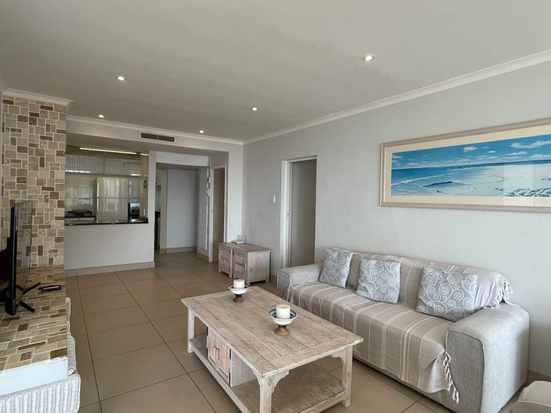 An open-plan living area featuring neutral tiled floors, recessed lighting, and a stone-clad feature pillar, seamlessly connecting to a functional kitchen space.