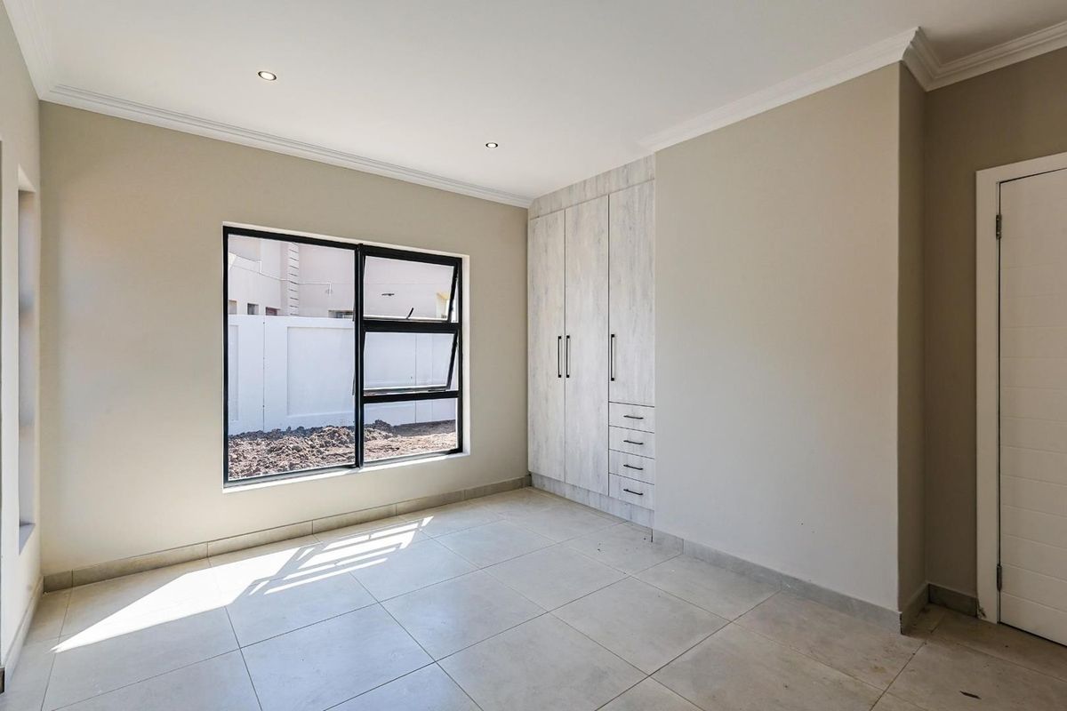 A bright, empty bedroom featuring neutral tiled flooring, modern built-in cupboards with a light wood finish, and large aluminum-framed windows.
