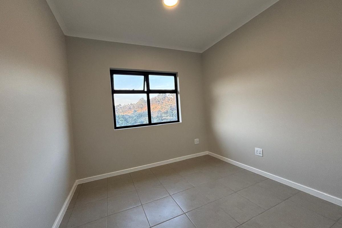 A clean, empty bedroom featuring neutral wall paint, tiled flooring, and a black aluminium window frame with a view of the surrounding landscape.