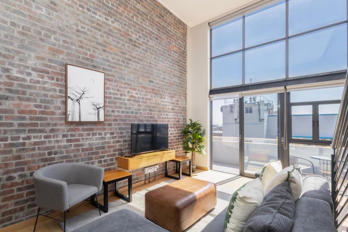 Spacious living room featuring a striking exposed brick feature wall, double-height windows, and direct access to a private balcony.
