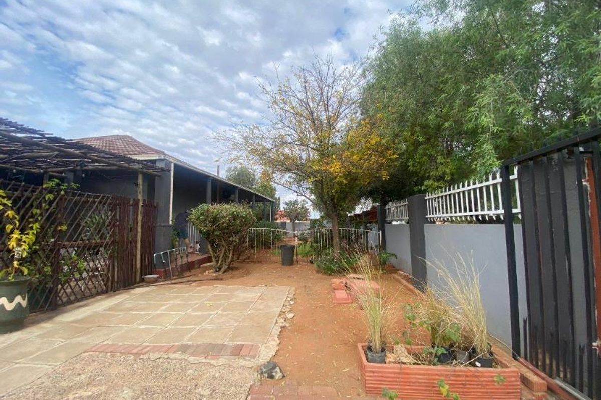 A rear garden area featuring a paved patio section, perimeter boundary walls with metal fencing, and established trees.