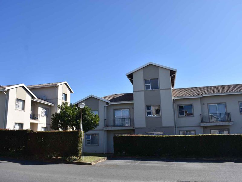 Exterior view of a multi-story apartment complex featuring tiled roofs, balconies with metal railings, and a paved parking area with landscaped hedging.