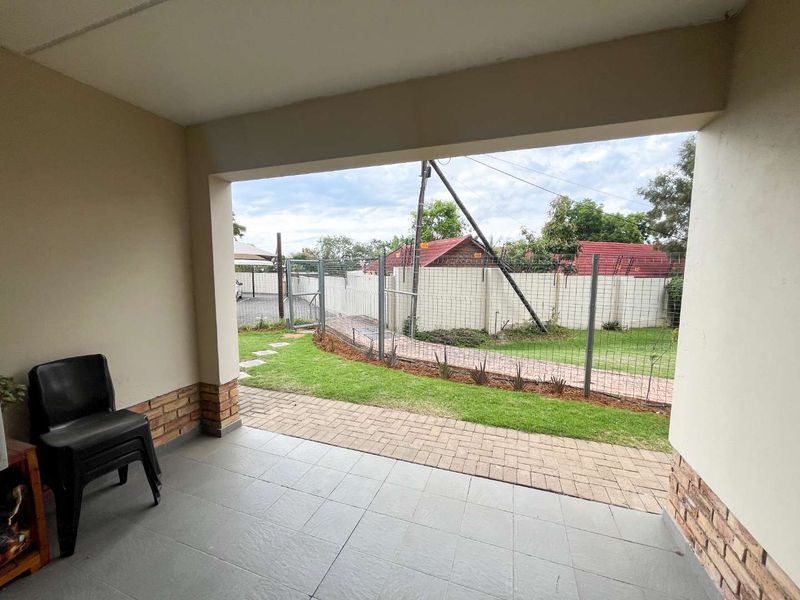 A covered patio area featuring tiled flooring and brick accents, overlooking a private garden space with a paved walkway and secure perimeter fencing.