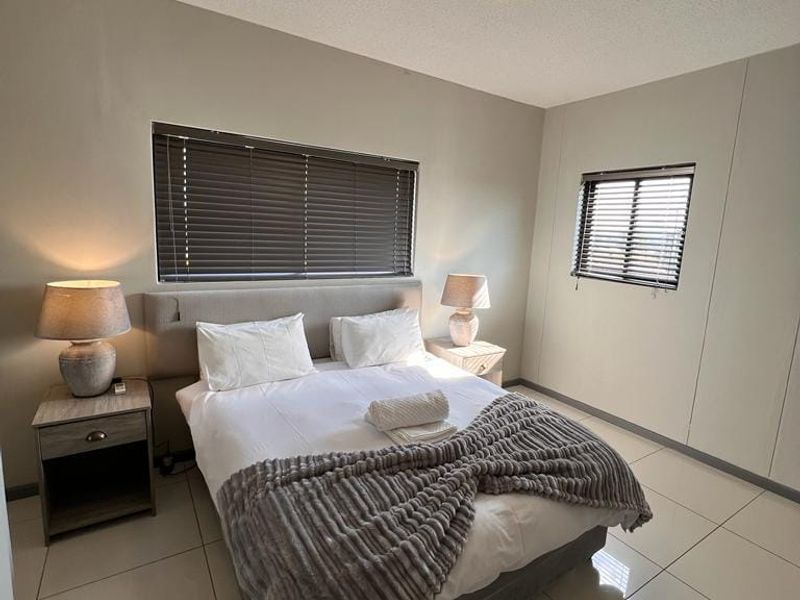 A well-maintained bedroom featuring neutral wall tones, large format floor tiles, and dual windows fitted with venetian blinds.