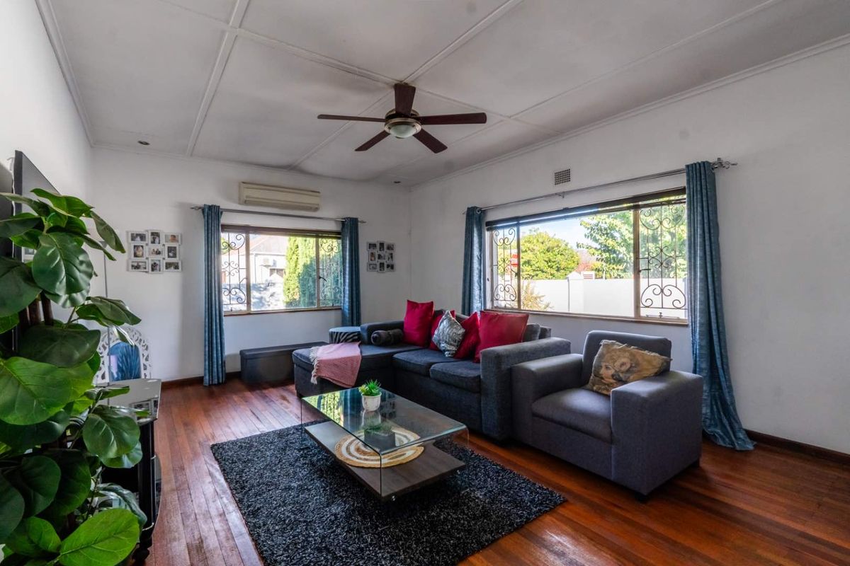 A comfortable living room featuring polished hardwood flooring, a ceiling fan, and split-system air conditioning, with large windows fitted with security bars.