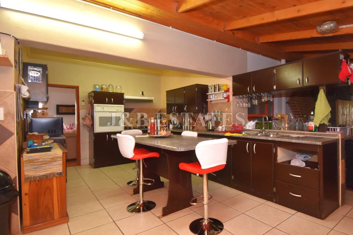 A functional kitchen featuring dark cabinetry, a central island with breakfast bar seating, tiled flooring, and exposed timber ceiling beams.