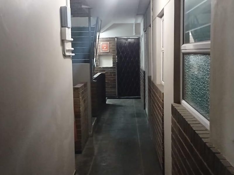 A narrow apartment hallway featuring brick wall accents, tiled flooring, and access to a staircase and security-gated entrance.