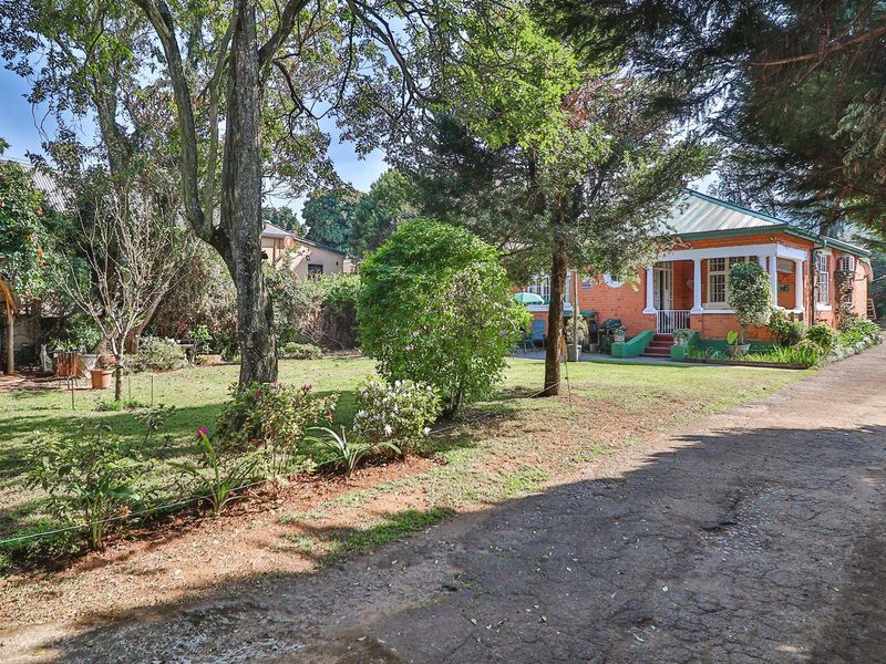 A charming brick house featuring a covered front porch, mature garden landscaping, and a long driveway, set within a spacious residential plot.