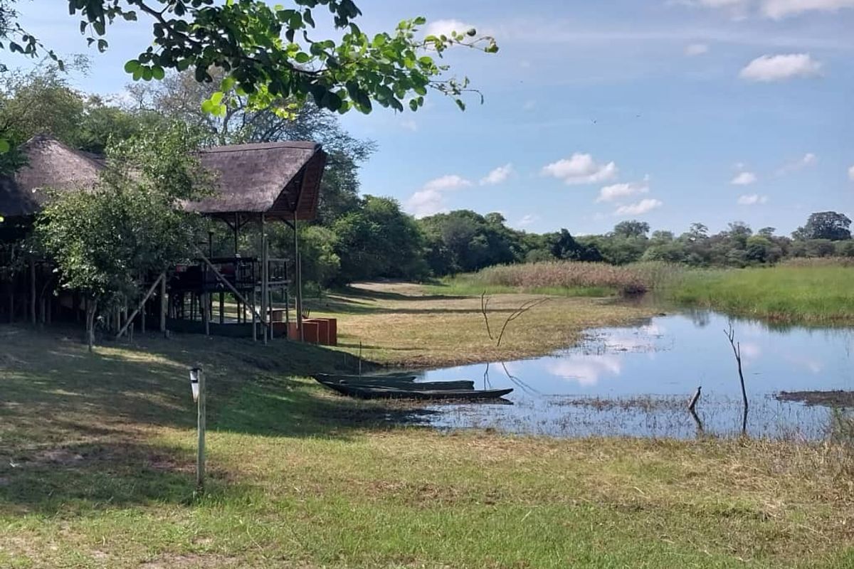 A scenic view of a lodge structure with a traditional thatched roof situated along the edge of a tranquil riverbank.