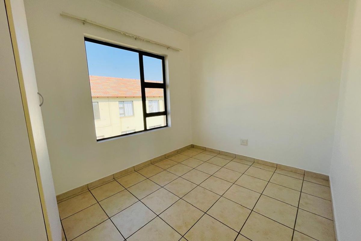 A bright, empty bedroom featuring neutral wall finishes, tiled flooring, and a large window providing natural light and a view of the complex.