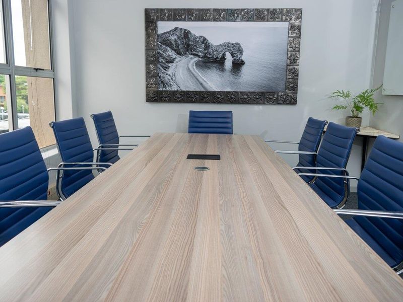 A professional office boardroom featuring a large central meeting table, carpeted flooring, and natural light from large windows.