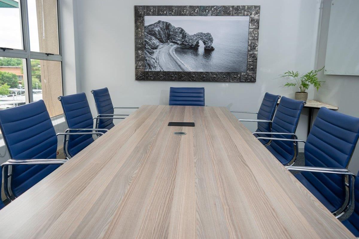 A professional office boardroom featuring a large central meeting table, carpeted flooring, and natural light from large windows.