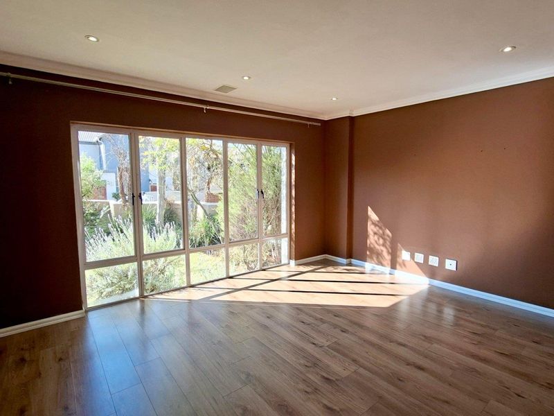 A bright, empty living room featuring modern laminate flooring, recessed downlighting, and large windows that provide ample natural light and garden views.