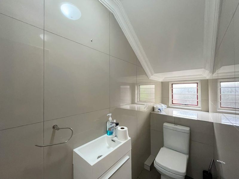 A compact, modern bathroom featuring floor-to-ceiling neutral tiling, a wall-mounted vanity basin, and a toilet, situated under a sloped ceiling.