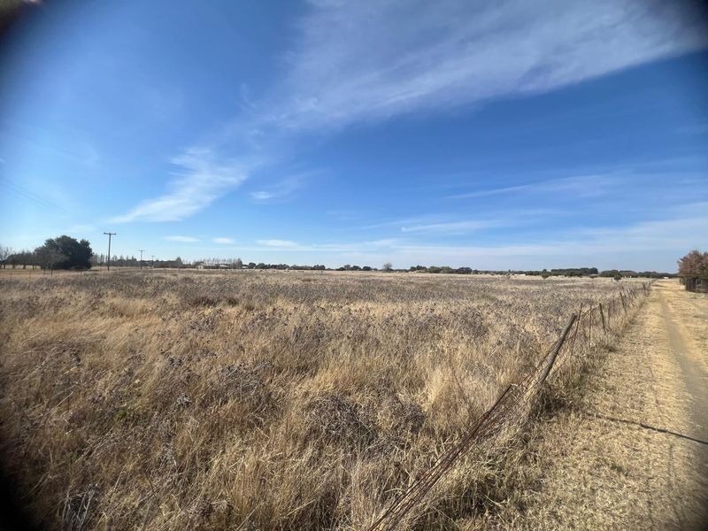 A wide-angle view of a vacant residential plot featuring dry grass, wire fencing, and an adjacent dirt access track under a clear blue sky.