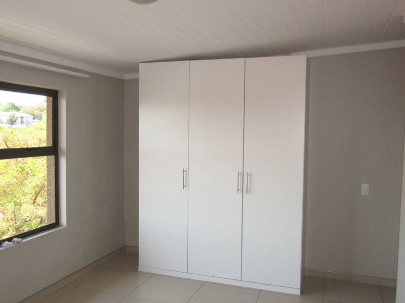 A bright, empty bedroom featuring neutral walls, tiled flooring, and a modern white built-in wardrobe with ample storage space.