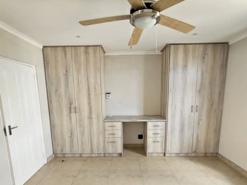 A clean bedroom featuring light wood-finish built-in cupboards, an integrated desk unit, a ceiling fan, and neutral tiled flooring.