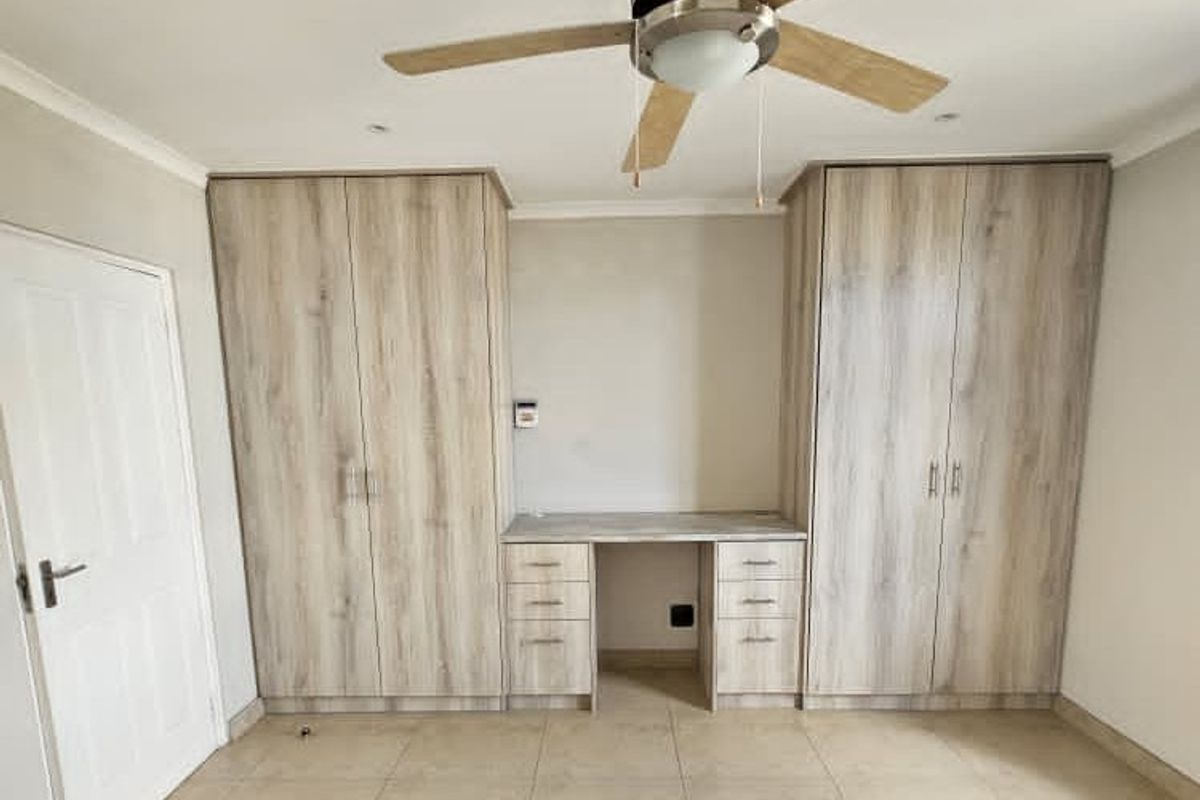 A clean bedroom featuring light wood-finish built-in cupboards, an integrated desk unit, a ceiling fan, and neutral tiled flooring.