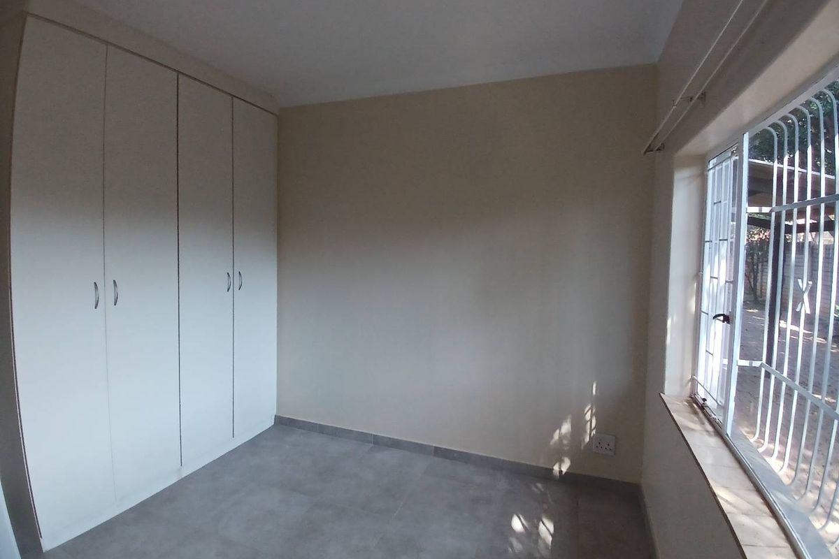 A clean, empty bedroom featuring neutral tiled flooring, built-in cupboards, and a window fitted with security bars.