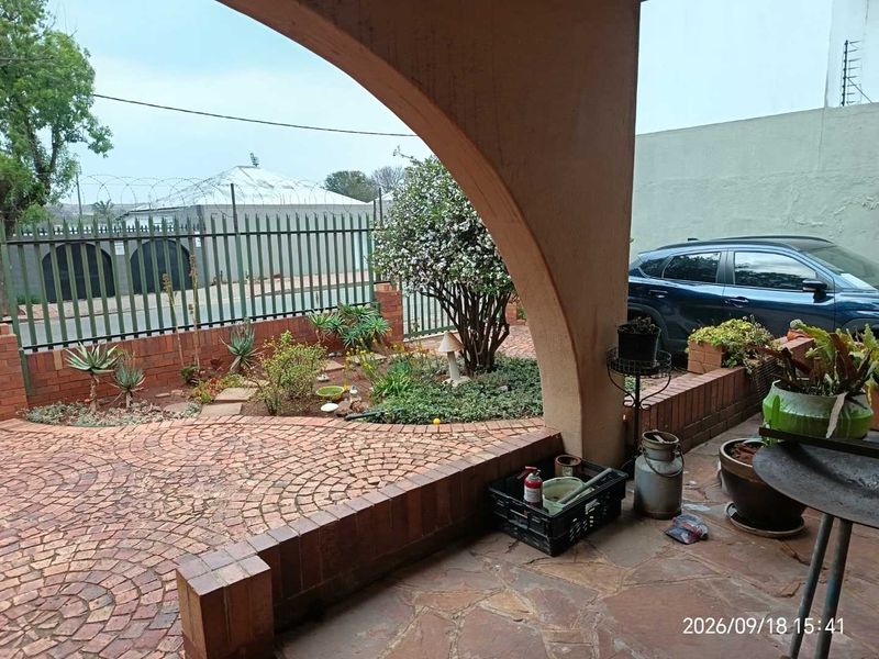A covered patio area featuring arched masonry, stone flooring, and brick paving, overlooking a landscaped garden with secure perimeter fencing.