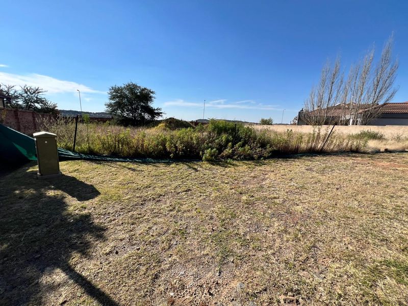 A view of a vacant residential plot featuring dry grass, overgrown vegetation, and a perimeter wall boundary.