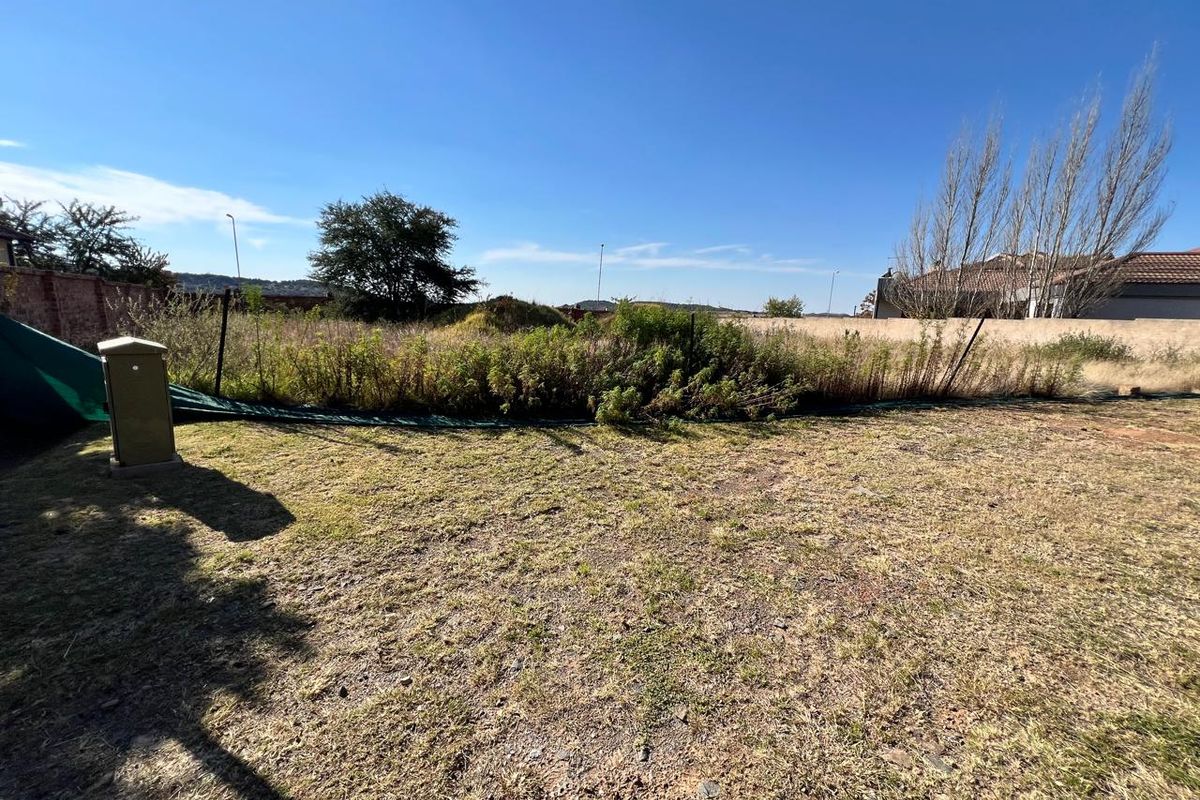 A view of a vacant residential plot featuring dry grass, overgrown vegetation, and a perimeter wall boundary.
