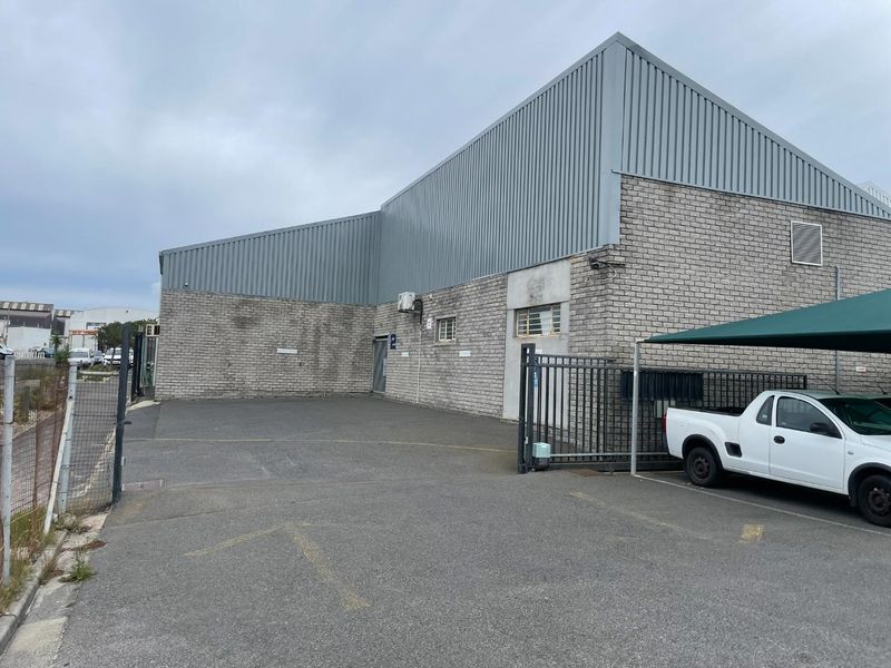 Industrial property exterior featuring concrete block construction, corrugated metal roofing, asphalt parking area, and a secure gated entrance with shade cloth carport.