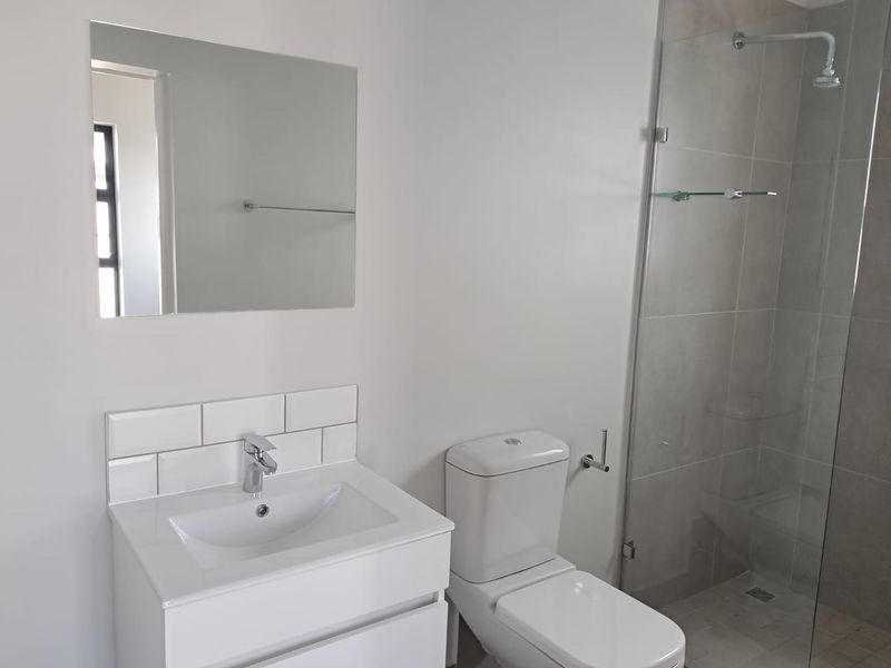 Modern bathroom featuring a sleek vanity with basin, wall-mounted mirror, toilet, and a glass-enclosed shower with contemporary tiling.