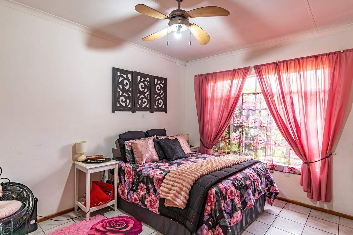 A bedroom featuring tiled flooring, a ceiling fan, and large windows with curtains, providing natural light and a functional living space.