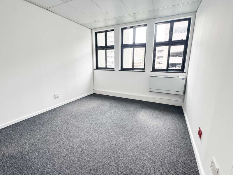 A clean, empty office space featuring grey carpet flooring, a suspended ceiling, and large windows providing natural light with an integrated HVAC unit.