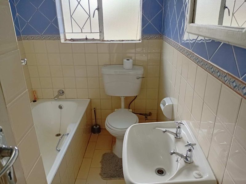 A dated bathroom featuring a standard bathtub, pedestal basin, and toilet with traditional wall tiling and security bars on the window.