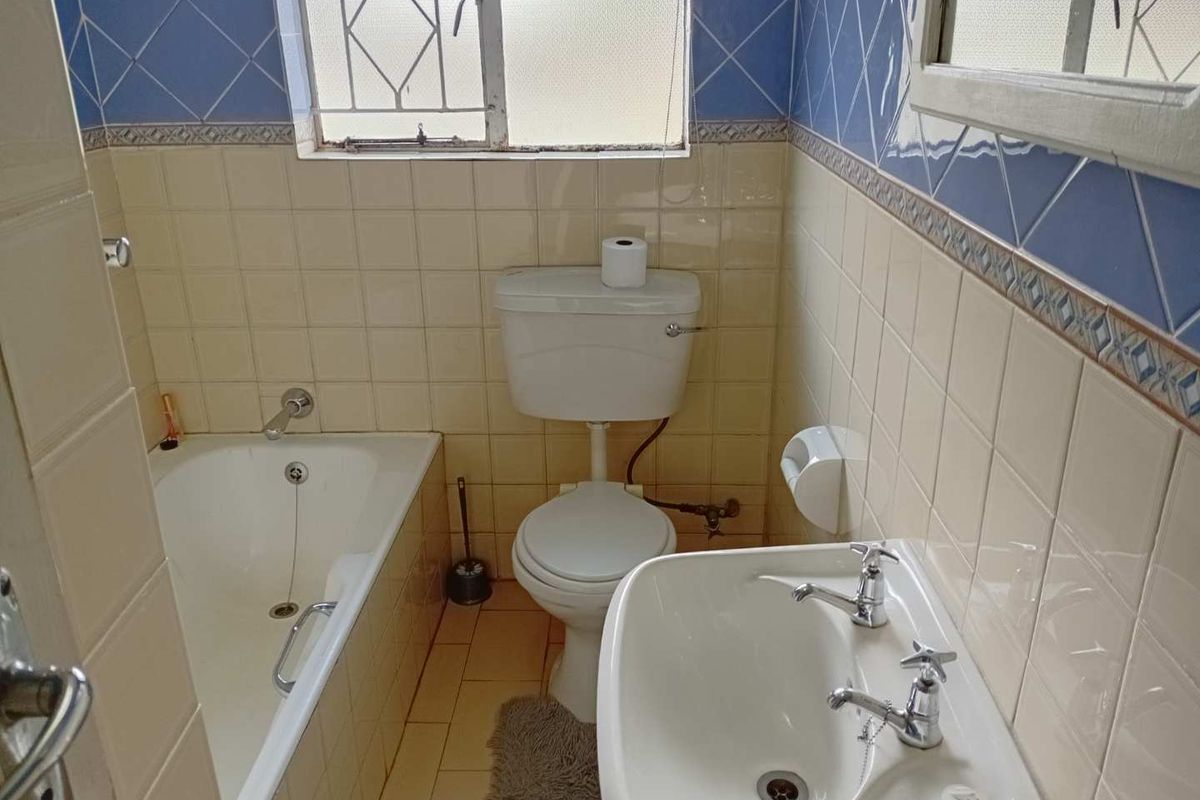 A dated bathroom featuring a standard bathtub, pedestal basin, and toilet with traditional wall tiling and security bars on the window.
