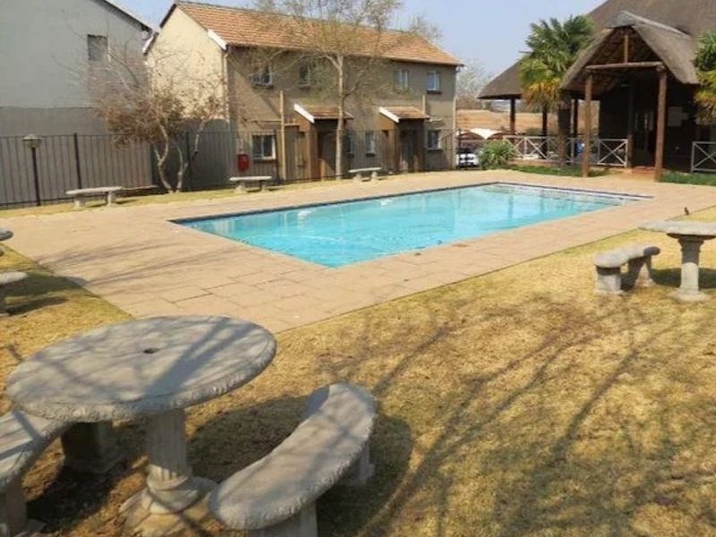 Communal swimming pool area featuring a paved deck, perimeter fencing, and adjacent thatched roof structure within a residential apartment complex.