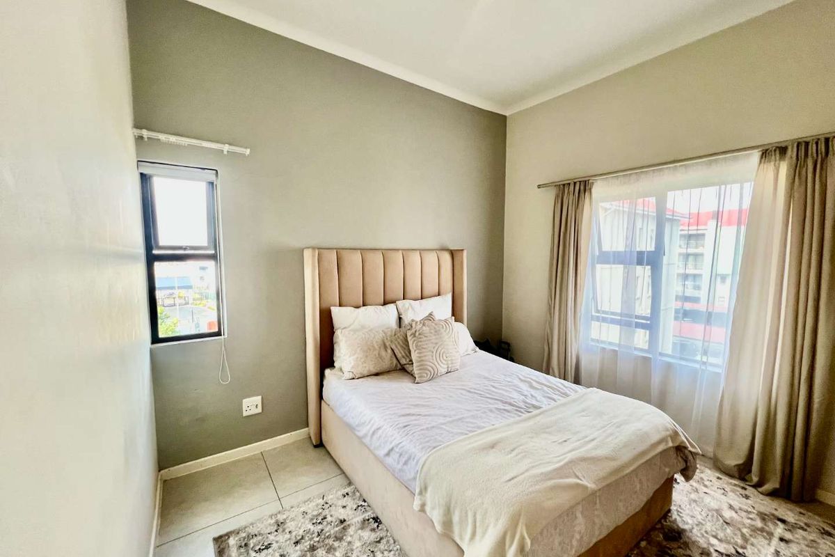 A well-lit bedroom featuring neutral wall tones, tiled flooring, and large windows that provide natural light and views of the surrounding apartment complex.