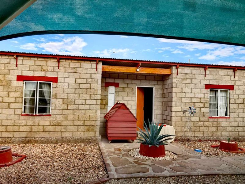 Rear exterior view of a concrete block farm building featuring a stone paved walkway, corrugated roof, and overhead shade sail protection.