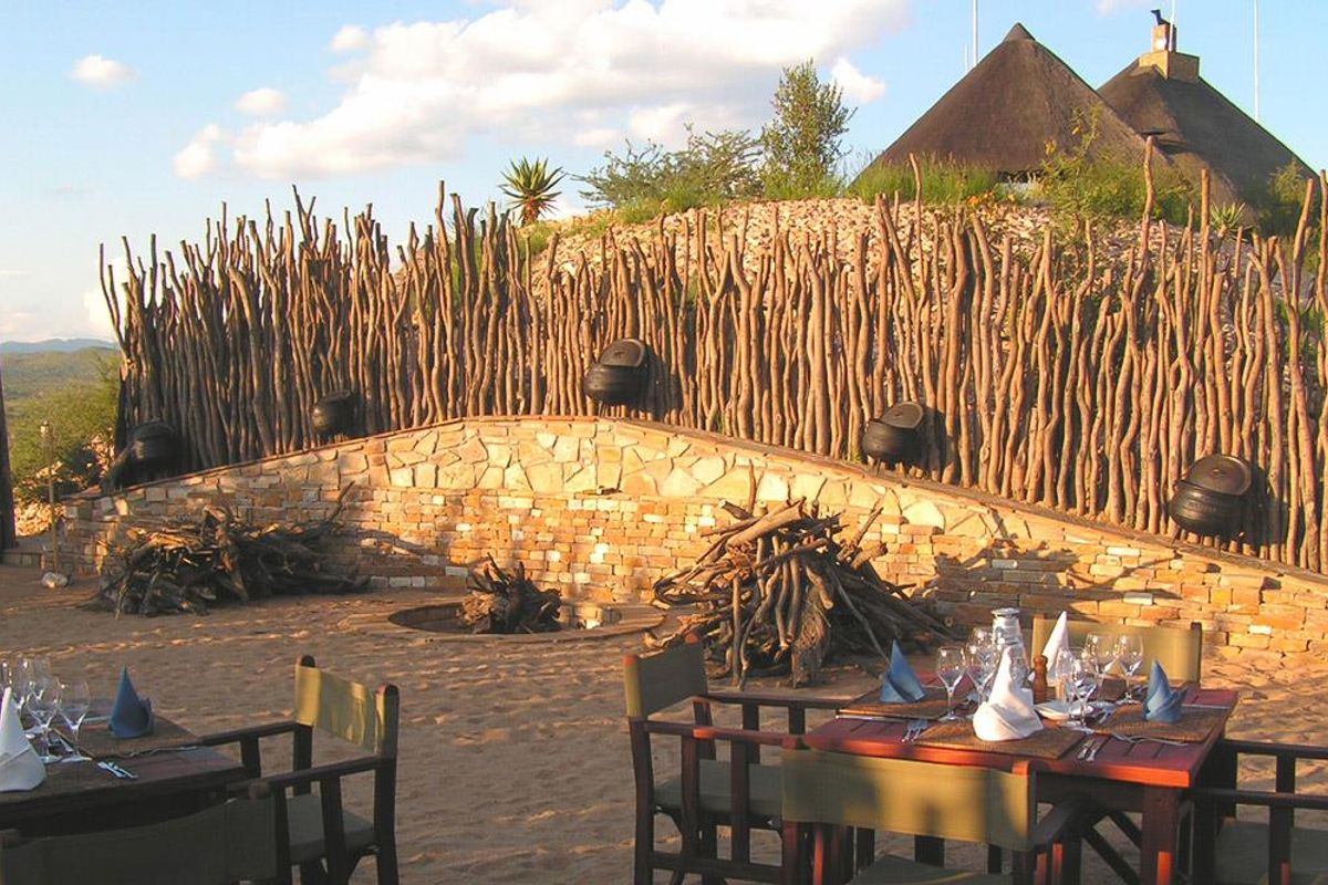 Outdoor entertainment area featuring a stone-walled fire pit, rustic branch fencing, and a sandy ground surface, set against a scenic farm landscape.