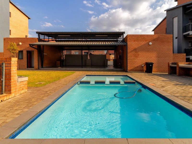 Communal outdoor area featuring a swimming pool with a shallow step section, paved surround, and a covered entertainment patio with a built-in braai.