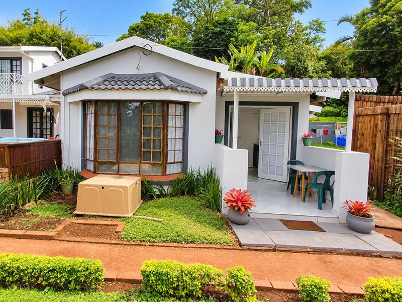 A rear exterior view of the apartment featuring a small tiled patio with a pergola, a bay window, and a landscaped garden area.
