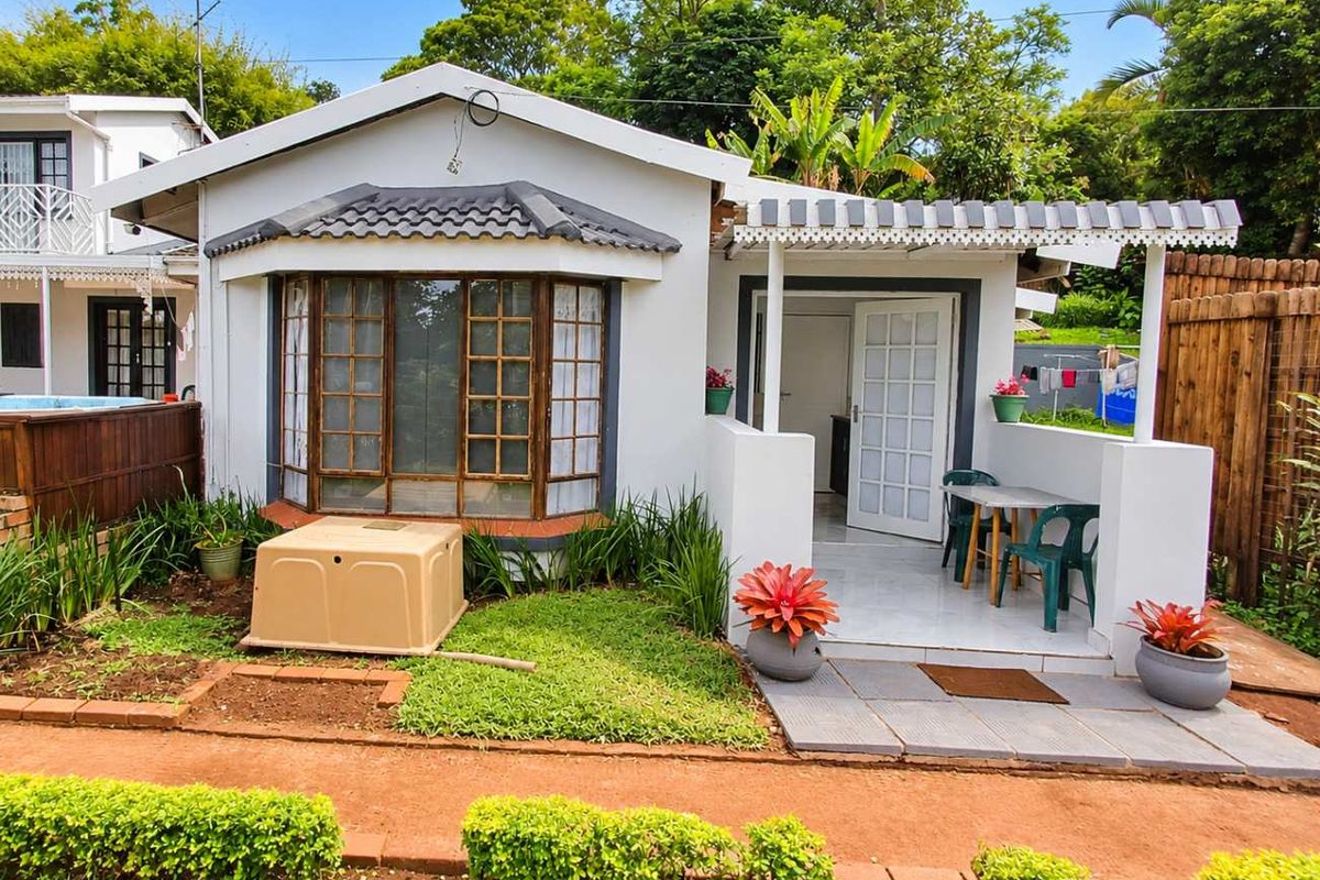 A rear exterior view of the apartment featuring a small tiled patio with a pergola, a bay window, and a landscaped garden area.