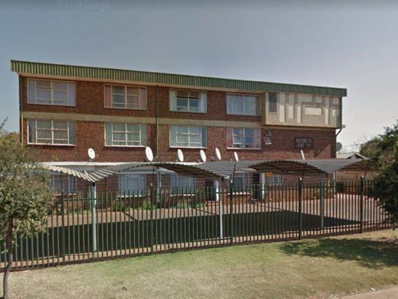 Front exterior view of a multi-story apartment building featuring brick facade, covered parking bays, and perimeter security fencing.