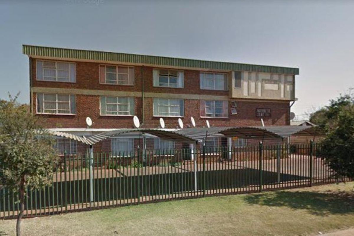 Front exterior view of a multi-story apartment building featuring brick facade, covered parking bays, and perimeter security fencing.