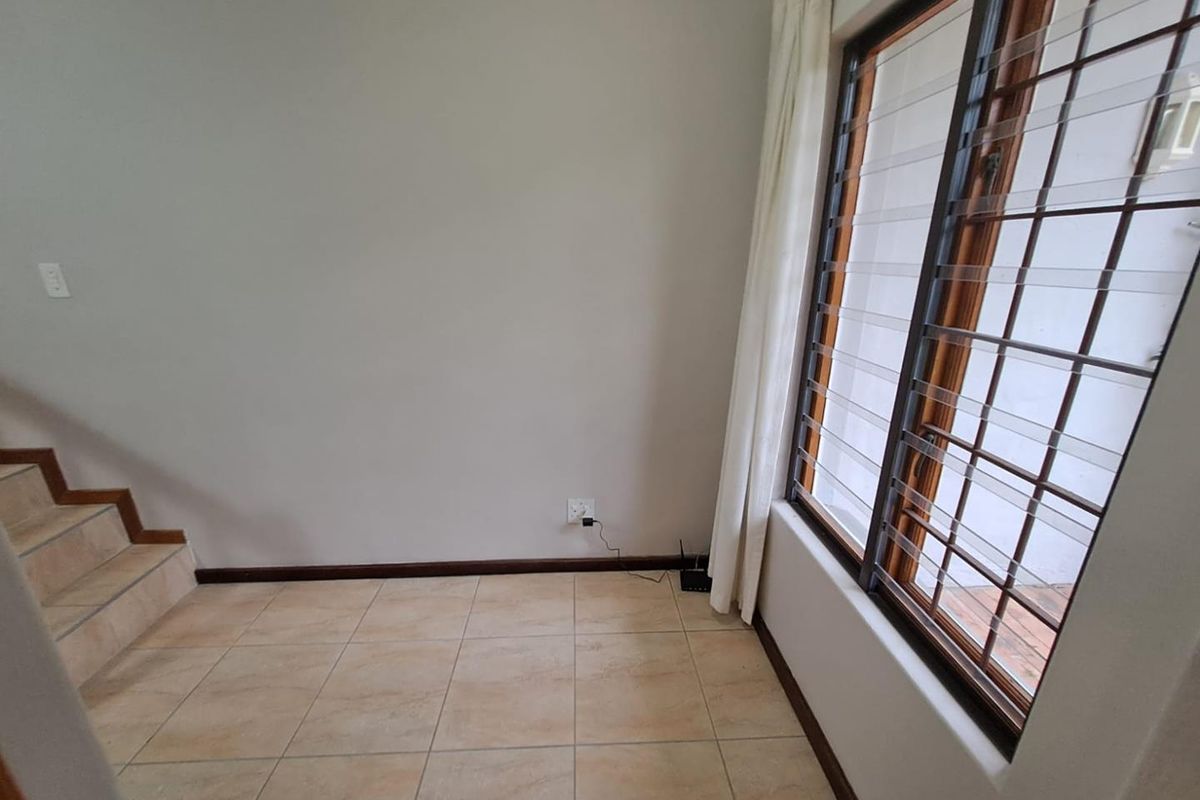 An empty living area featuring tiled flooring, a staircase, and large windows with security bars, offering a bright and functional space.