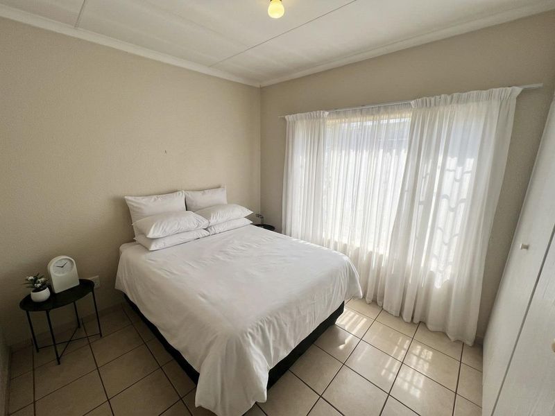 A functional bedroom featuring neutral tiled flooring, a built-in wardrobe, and large windows dressed with sheer curtains, providing a bright and simple living space.