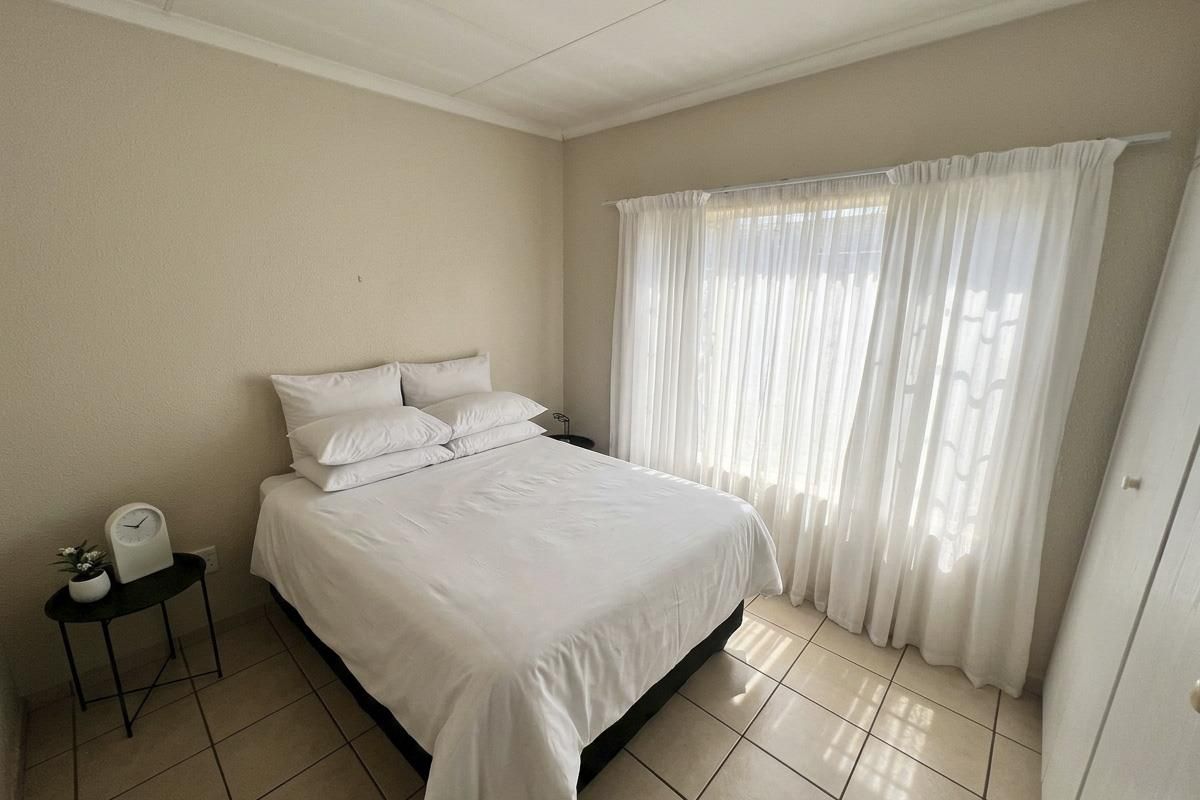 A functional bedroom featuring neutral tiled flooring, a built-in wardrobe, and large windows dressed with sheer curtains, providing a bright and simple living space.