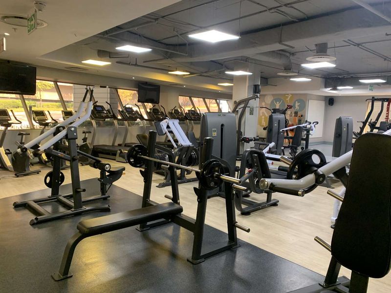 A well-equipped commercial gym facility featuring industrial-style exposed ceilings, recessed LED lighting, rubberized flooring, and large windows providing natural light.