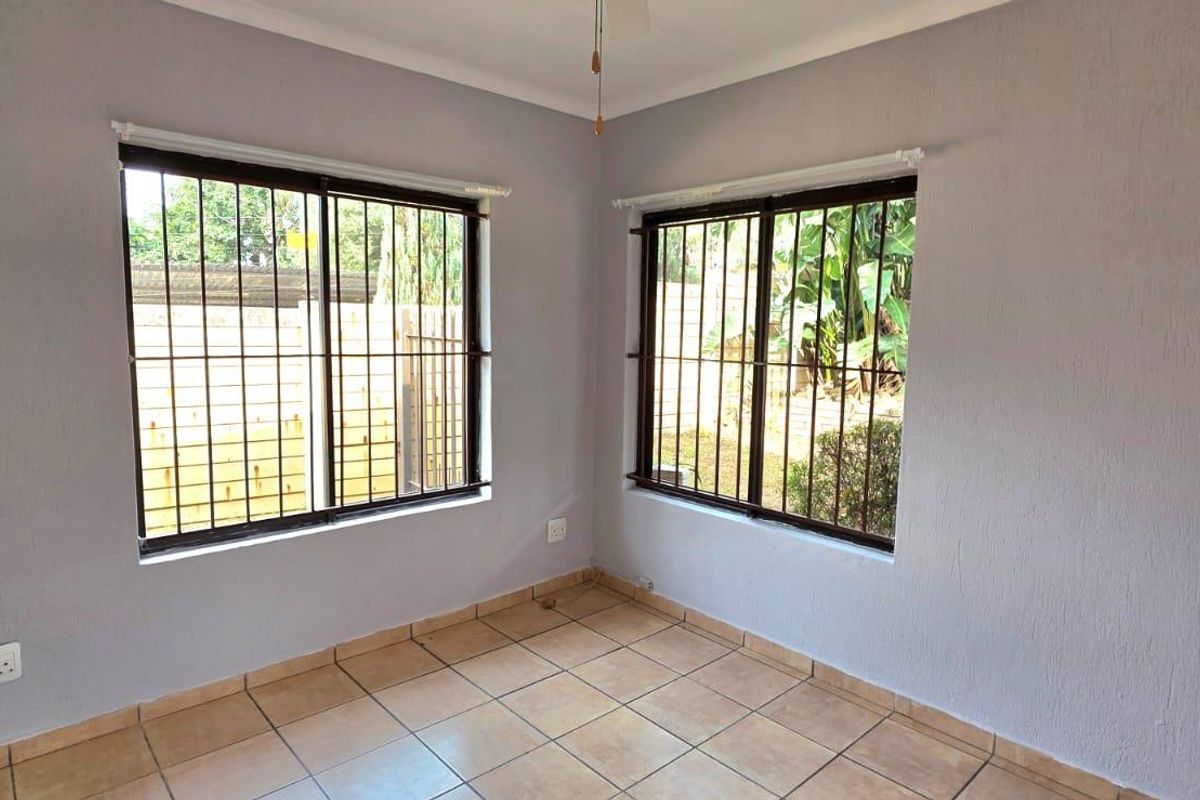 A bright, empty bedroom featuring neutral grey walls, tiled flooring, and large windows fitted with security bars.