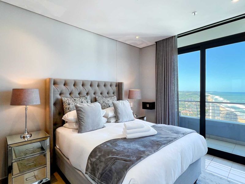 Modern bedroom featuring neutral finishes, recessed lighting, and large sliding glass doors opening to a balcony with expansive coastal views.